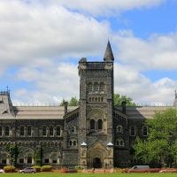 何一苇在郑州大学校园里走访记录生活细节的随手拍，手里拿着笔记本，背景是树荫下的步道与教学楼一角